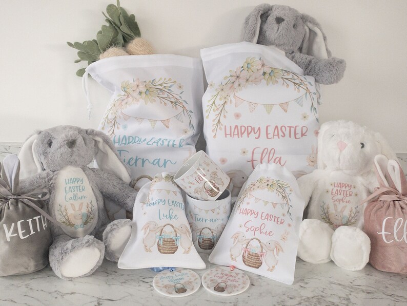 K&ouml;nnte beinhalten: Eine Auswahl an Ostergeschenken, darunter Pl&uuml;schhasen, Kordelzugbeutel und Tassen. Die Beutel und Tassen sind mit Blumen- und Wimpeldesigns, dem Text "Frohe Ostern" und Namen verziert. Die Hasen sind grau und wei&szlig;.