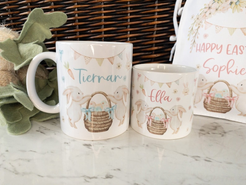 K&ouml;nnte beinhalten: Wei&szlig;er Keramikbecher und Tasse mit Ostermotiv. Die Designs zeigen Hasen, Osterk&ouml;rbe und die Namen "Tiernan" und "Ella". Ein passender Kordelzugbeutel mit "Happy Easter Sophie" ist ebenfalls zu sehen.