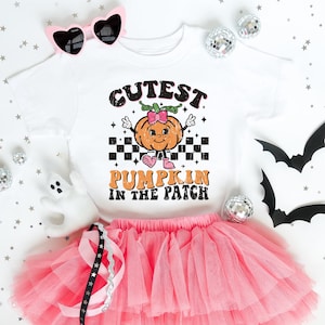 Può includere: Una t-shirt bianca a maniche corte con la grafica di una zucca arancione con un fiocco rosa, stivali rosa e segni di pace. Il testo recita CUTEST PUMPKIN IN THE PATCH. La t-shirt è abbinata a una gonna in tulle rosa a strati.