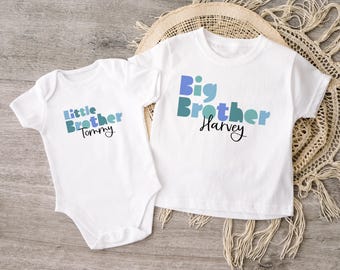 Puede incluir: Dos camisetas blancas para bebés. Una es un body con "Little Brother Tommy" en letras azules y turquesas. La otra es una camiseta con "Big Brother Harvey" en letras azules y turquesas. Ambas camisetas están sobre una superficie de madera.