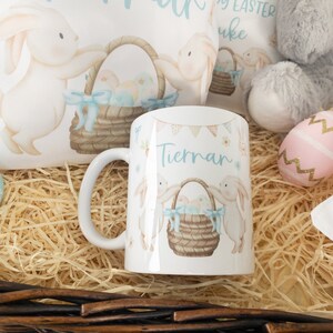 K&ouml;nnte beinhalten: Wei&szlig;e Keramiktasse mit Osterhasen-Design, einem Korb mit Eiern und dem Namen "Tiernan" in Hellblau. Die Tasse ist von Osterdekorationen umgeben, darunter ein Pl&uuml;schhase und dekorative Eier.