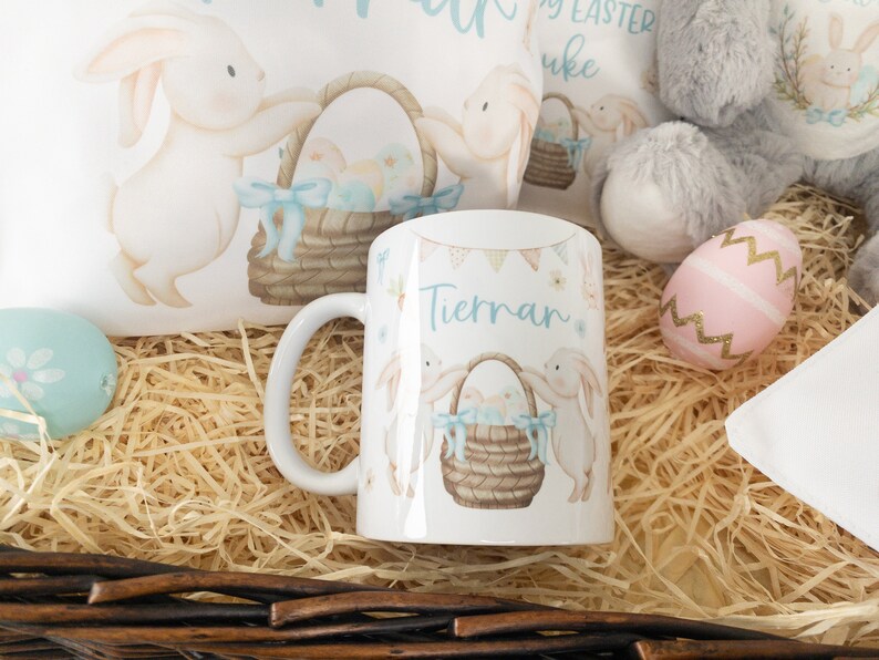 K&ouml;nnte beinhalten: Wei&szlig;e Keramiktasse mit Osterhasen-Design, einem Korb mit Eiern und dem Namen "Tiernan" in Hellblau. Die Tasse ist von Osterdekorationen umgeben, darunter ein Pl&uuml;schhase und dekorative Eier.