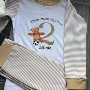 May include: Children's white and beige striped pyjamas featuring a teddy bear graphic and the text "When I wake up, I'll be 2 Lennie". The pyjamas have beige trim and a soccer ball graphic.