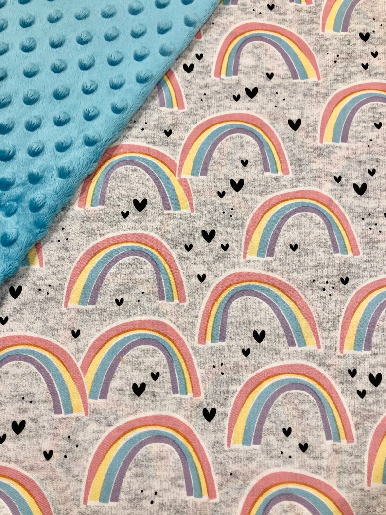 Puede incluir: Una manta para beb&eacute; con tela de lunares minky azul claro y una tela gris estampada con arco&iacute;ris pastel y corazones negros. Los arco&iacute;ris tienen arcos rosas, amarillos, azules y morados. La manta es suave.