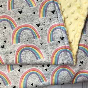 Puede incluir: Una manta gris con un patr&oacute;n repetido de coloridos arco&iacute;ris y peque&ntilde;os corazones negros. La manta est&aacute; doblada, revelando un reverso suave, amarillo y texturizado.