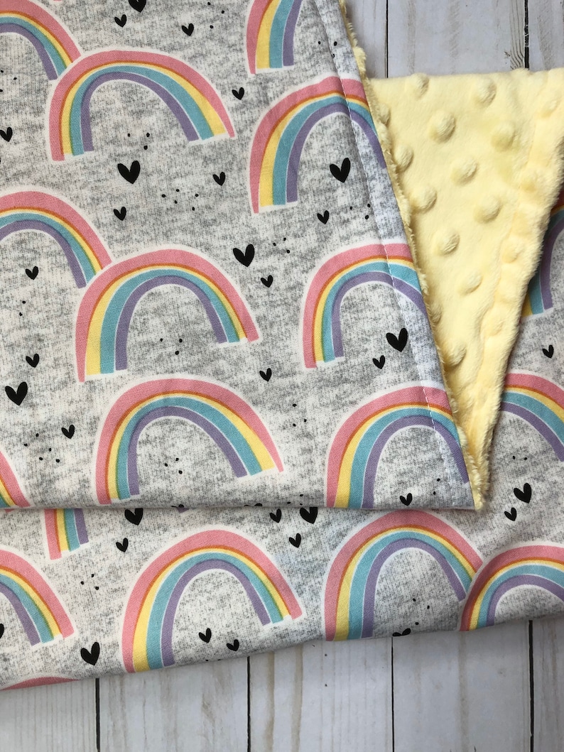 Puede incluir: Una manta gris con un patr&oacute;n repetido de coloridos arco&iacute;ris y peque&ntilde;os corazones negros. La manta est&aacute; doblada, revelando un reverso suave, amarillo y texturizado.