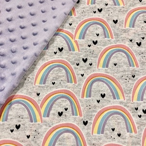 Puede incluir: Una manta para beb&eacute; con un fondo gris con coloridos arco&iacute;ris y corazones negros. La manta tiene una tela minky lavanda suave y texturizada en el lado izquierdo. Los arco&iacute;ris tienen rayas rosas, amarillas, azules y moradas.
