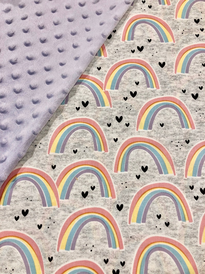 Puede incluir: Una manta para beb&eacute; con un fondo gris con coloridos arco&iacute;ris y corazones negros. La manta tiene una tela minky lavanda suave y texturizada en el lado izquierdo. Los arco&iacute;ris tienen rayas rosas, amarillas, azules y moradas.