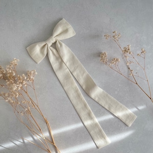 Könnte beinhalten: Eine cremefarbene Schleife aus Leinen mit langen, fließenden Bändern. Die Schleife ist in der Mitte gebunden und hat eine klassische Form. Der Stoff hat eine subtile Textur. Trockenblumen sind um die Schleife auf einer hellgrauen Oberfläche angeordnet.