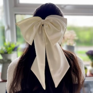 Peut inclure: Un grand nœud en satin blanc noué dans les cheveux d'une personne. Le nœud est noué en un grand nœud lâche et les extrémités du nœud sont longues et flottantes.