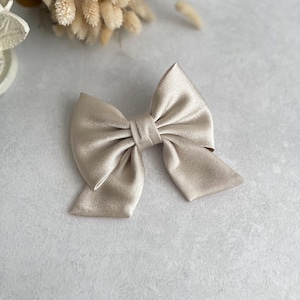 May include: A large, beige satin bow hair clip. The bow is tied in a classic shape with two loops and a center knot.