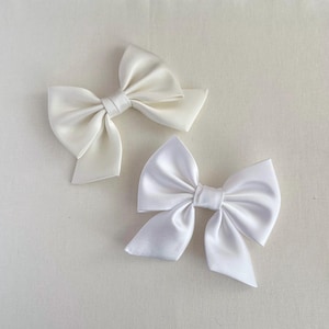 May include: Two hair bows made of smooth, lustrous fabric. One bow is a soft cream colour, while the other is a bright, crisp white. Both bows feature a classic tied design with wide, structured loops and short, angled tails extending from the centre.