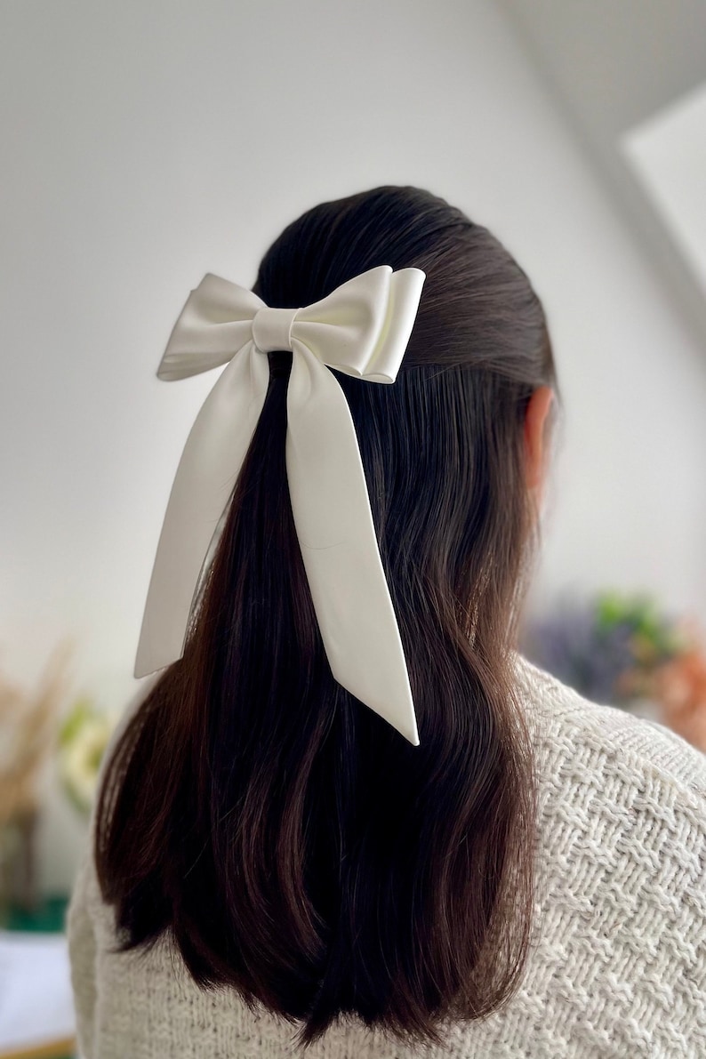 Puede incluir: Un lazo de sat&eacute;n blanco con colas largas, atado en el cabello de una persona.