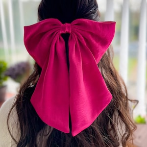 May include: A large, vibrant fuchsia bow hair accessory. The bow is made of a soft fabric and is tied in the centre, with long, flowing tails. The bow is the focal point, set against a blurred background. The hair accessory adds a pop of colour.