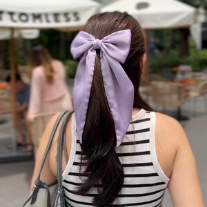 Könnte beinhalten: Ein helllila Haarschmuck mit Schleife, in einem Pferdeschwanz gebunden. Die Schleife ist aus einem weichen, fließenden Stoff gefertigt. Die Person trägt ein schwarz-weiß gestreiftes Tanktop.