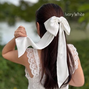 Puede incluir: Lazo para el pelo de satén blanco marfil con largas cintas. El lazo está atado en la parte posterior de la cabeza, con el texto "Ivory White" en la esquina superior derecha. La persona lleva un top de encaje blanco.