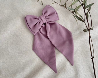May include: A large, lavender-colored hair bow with long, flowing tails. The bow is made of a smooth fabric and is centered on a neutral-toned background. A sprig of dried greenery is placed to the side of the bow.