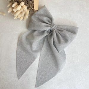 Könnte beinhalten: Eine große silberne Glitzer Schleife mit langen Bändern. Die Schleife ist zu einem Knoten gebunden und die Bänder fließen.
