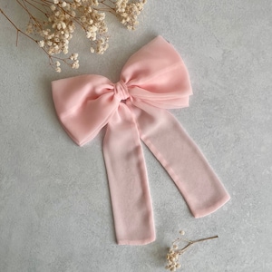 Könnte beinhalten: Eine große rosafarbene Schleife mit langen Bändern. Die Schleife ist aus einem weichen, durchsichtigen Stoff gefertigt und perfekt, um jedem Outfit einen Hauch von Eleganz zu verleihen.