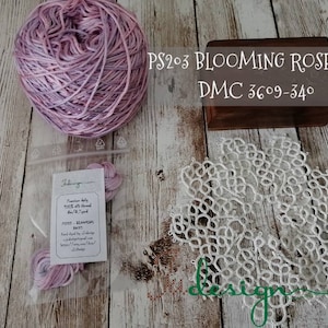 Può includere: Un gomitolo di filo di seta rosa e viola etichettato "PS203 BLOOMING ROSE silk DMC 3609-340" insieme a un pizzo bianco e una piccola borsa di filo.