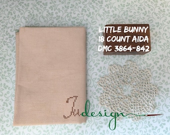 Peut inclure: Un morceau plié de tissu Aida beige avec une initiale brodée, une boîte en bois avec le texte "LITTLE BUNNY 18 COUNT AIDA DMC 3864-842", et un napperon en dentelle blanche. Les articles sont disposés sur un fond à motifs.