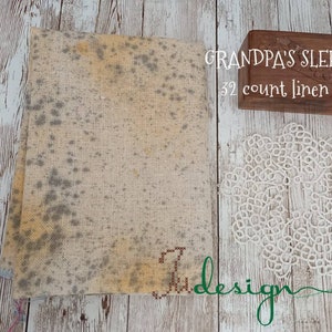 Peut inclure: Un morceau plié de tissu de lin beige avec des marques grises et jaunes, une boîte en bois avec le texte "GRANDPA'S SLEEVE 32 count linen", et un napperon en dentelle blanche. Le mot "design" est écrit en vert.