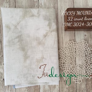 Op de afbeelding: Een stuk crèmekleurig linnen stof met een geborduurde bruine letter "J" in de rechterbenedenhoek. De stof is gelabeld met "Rocky Mountain 32 count linen DMC 3024-3032".