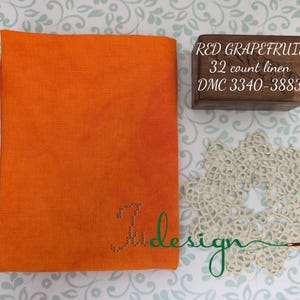 May include: An orange fabric square with the embroidered word "Judesign" in green. A small wooden box with the text "RED GRAPEFRUIT 32 count linen DMC 3340-3883" is next to the fabric. A cream-colored doily is also in the image.