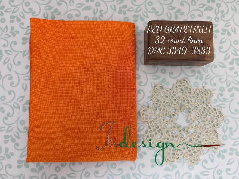 May include: An orange fabric square with the embroidered word "Judesign" in green. A small wooden box with the text "RED GRAPEFRUIT 32 count linen DMC 3340-3883" is next to the fabric. A cream-colored doily is also in the image.