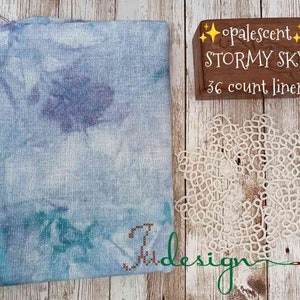 May include: A piece of blue and green linen fabric with a white background. The fabric is labeled "Opalescent Stormy Sky 36 count linen".