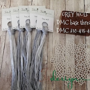 Könnte beinhalten: Fünf Knäuel grauer Stickgarn mit Etiketten mit der Aufschrift "Grey Wolf" und "DMC 8/6.7". Ein Holzblock mit dem Text "GREY WOLF DMC base thread DMC 318-415-414" und eine weiße Spitzendeckchen.