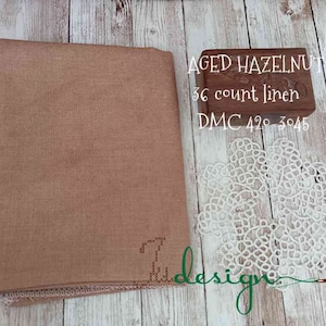 May include: Aged hazelnut 36 count linen fabric with a white lace doily and a small wooden box. The box has the text "AGED HAZELNUT" and "DMC 420-3045". The fabric is folded and has a textured appearance.