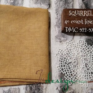 May include: A rectangular piece of mustard-coloured linen fabric with a visible weave, accompanied by a dark wooden block labelled SQUIRREL 40 count linen DMC 922-976 and a white circular lace doily featuring an intricate, knotted, floral-like pattern.
