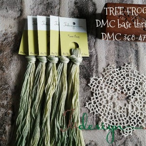 May include: Five skeins of green embroidery floss with labels that say "Tree Frog" and "DMC base thread". A wooden box with the text "TREE FROG DMC base thread DMC 368-472" and a white crocheted doily.