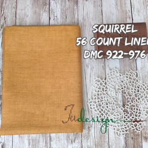 May include: A rectangular piece of mustard yellow 56 count linen fabric with a small cross-stitched logo in the corner. Next to it is a white lace doily and a wooden block with the text SQUIRREL 56 COUNT LINEN DMC 922-976 printed on it.