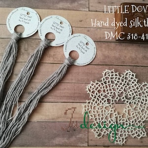 May include: Three bundles of Little Dove hand-dyed silk thread in gray, each attached to a circular tag. The tags have text. A white, lace doily is in the lower right. The text "LITTLE DOVE Hand dyed silk thread DMC 318-415" is in the upper right.