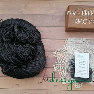 Puede incluir: Un ovillo de hilo negro está junto a una pequeña caja de madera con el texto "PS98 - EBEN silk DMC 310". Un paquete de hilo negro está dentro de una bolsa de plástico transparente, colocado sobre un tapete de encaje blanco. La palabra "Jadesign" es visible.