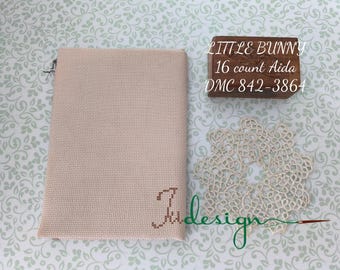 Peut inclure: Tissu beige pour point de croix avec la lettre "J" brodée en marron. Une boîte en bois avec le texte "LITTLE BUNNY 16 count Aida DMC 842-3864" est visible. Un napperon crème et le mot "Judesign" sont également présents.