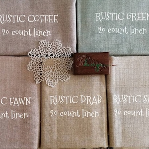 Könnte beinhalten: Eine Auswahl an Leinenstoffmustern in gedeckten Farben: Kaffee, Grün, Rehbraun, Schlamm und Schaf. Jedes Muster ist mit seinem Farbnamen und "20 count linen" in weißer Schrift beschriftet. Eine weiße Spitzendeckchen und ein Holzakzent sind ebenfalls vorhanden.