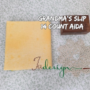 Può includere: Un tessuto Aida giallo da 14 punti con la scritta "Grandma's Slip" e un'iniziale ricamata a punto croce. Sono presenti anche una piccola scatola di legno e un centrino di pizzo color crema.
