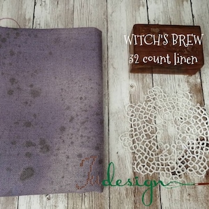 May include: A folded piece of lavender linen fabric with dark gray spots, a small wooden box with the text "WITCH'S BREW 32 count linen", and a white lace doily. The fabric has a textured appearance, and the doily has an intricate, openwork design.