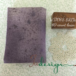 May include: A folded piece of purple fabric with a mottled pattern, a small wooden box with the text "WITCH'S BREW 40 count linen", and a cream-coloured lace doily. The items are arranged on a patterned background.