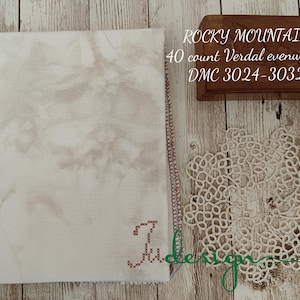May include: A cream-colored fabric with a mottled pattern, a decorative doily, and a wooden box with text. The fabric has a stitched edge and a cross-stitched initial. The text on the box reads "Rocky Mountain".