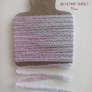 Può includere: Un rotolo di nastro di pizzo color lampone, largo 10 mm, è esposto su un tagliere di legno. Il nastro è bianco delicato con un effetto ombre rosa. Il testo "RASPBERRY PARFAIT" e "10mm" sono visibili.