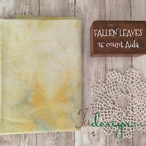 Puede incluir: Tela Aida con tinte amarillo y naranja, una caja de madera con el texto "FALLEN LEAVES 16 count Aida" y un tapete de encaje blanco. Los artículos están dispuestos sobre una superficie de madera.
