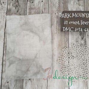 Op de afbeelding: Een lichtgrijze linnen stof, een wit kanten kleedje en een houten bord met de tekst "DARK MOUNTAIN 32 count linen DMC 3024-648" zijn gerangschikt op een houten oppervlak. Het kleedje heeft een bloemenpatroon.