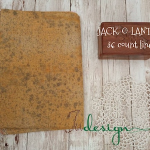 Puede incluir: Un trozo de tela de lino de 36 hilos, de color marrón y amarillo, con un patrón moteado. La tela está doblada y etiquetada con "JACK-O-LANTERN 36 count linen".