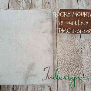 Op de afbeelding: Een witte linnen stof, een houten doos met de tekst "ROCKY MOUNTAIN 28 count linen DMC 3024-3032" en een wit kanten kleedje worden gepresenteerd op een houten oppervlak. De stof heeft een subtiel patroon en het kleedje heeft een ingewikkeld ontwerp.