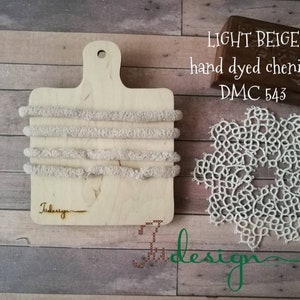 Puede incluir: Hilo de chenilla teñido a mano beige claro en una tabla de madera con el texto "Tiddesign" y una etiqueta DMC 543. Un encaje de ganchillo blanco está sobre la superficie de madera.