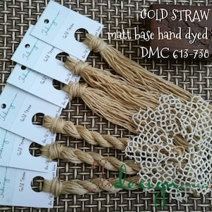Puede incluir: Seis carretes de hilo de bordar teñido a mano de color beige claro. Los carretes están etiquetados con "Gold Straw" y "6/87" con la marca "Fidesign". El hilo se muestra sobre una superficie tejida marrón con un encaje blanco y un letrero de madera que dice "GOLD STRAW matt base hand dyed thread DMC 613-738".